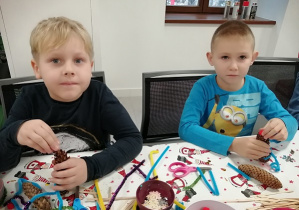 Chłopcy siedzą przy stole i przyklejają elementy na szyszkę